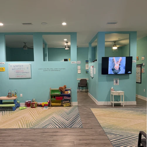 A wide view of a clinic interior with light blue walls, children’s toys, chairs, rugs, and a wall-mounted screen.