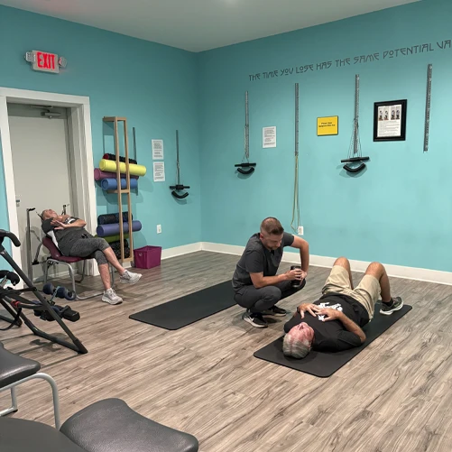 Two adults on exercise mats in a clinic room, one lying down and one kneeling beside, with equipment