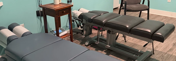 Two padded chiropractic adjustment tables positioned in a clinic room with chairs, small tables, and wall art.