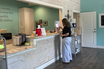 A person standing at a reception counter inside a clinic, with shelves, paperwork, and office items visible.