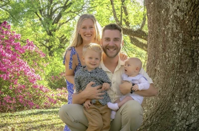 Two adults and two children seated outdoors near a large tree, surrounded by greenery and flowering plants.