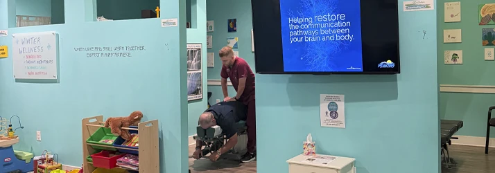 A clinic room with a screen on the wall, toys on shelves, and a person adjusting another on a padded table.
