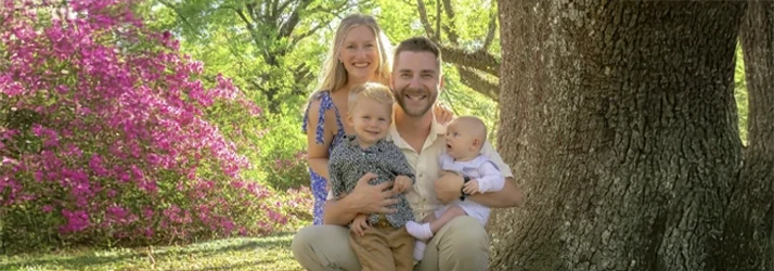 Dr. Christian Reagan, his wife and two children seated outdoors near a large tree, surrounded by greenery and flowering plants.